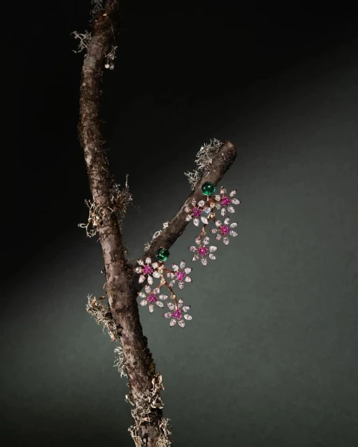 Elegant floral-themed earrings with rubies, diamonds, and emeralds, showcased on a moss-covered branch against a dark background.