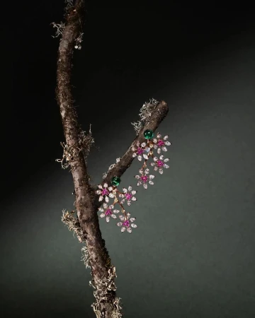Elegant floral-themed earrings with rubies, diamonds, and emeralds, showcased on a moss-covered branch against a dark background.