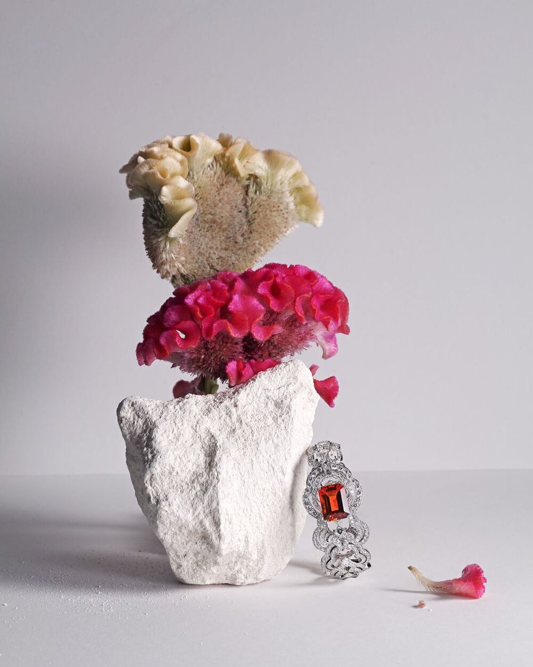 Still life featuring a diamond bracelet with an orange gemstone, white rock, and pink and white celosia flowers against a white background.