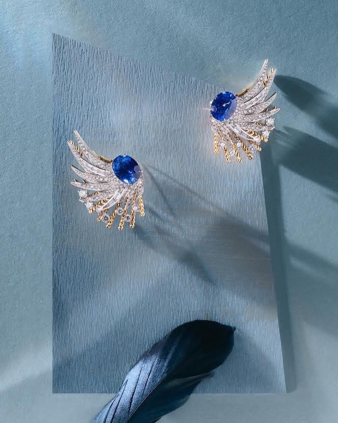 Elegant pair of blue sapphire and diamond wing-shaped earrings on a light blue textured paper background with a dark feather.