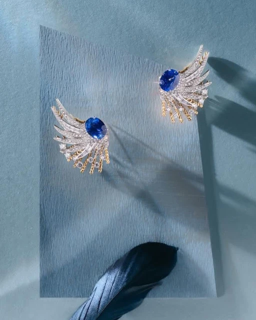 Elegant pair of blue sapphire and diamond wing-shaped earrings on a light blue textured paper background with a dark feather.