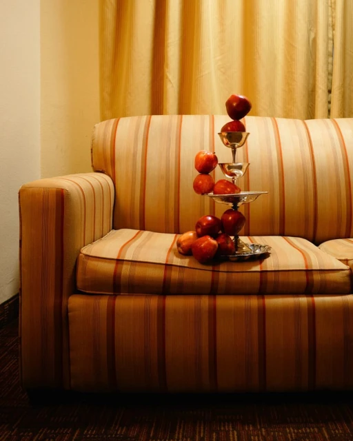 A striped couch has a silver tiered server stacked with red apples in an indoor setting with a tan striped curtain in the background.