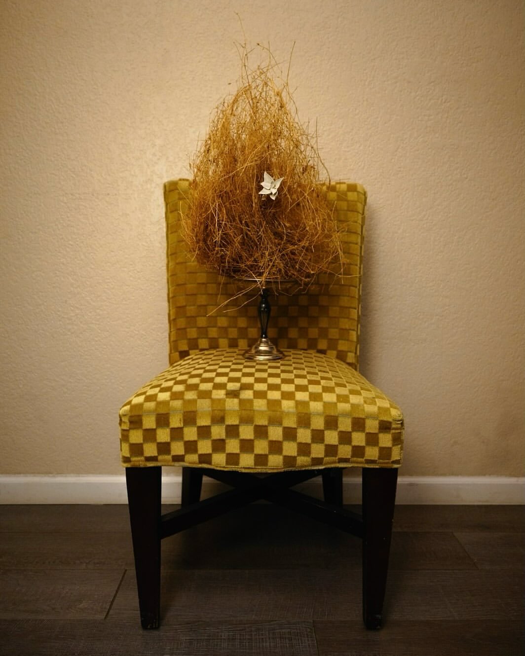 Still life photo of a yellow checkerboard chair with dried floral arrangement in a vintage metal candle holder against a cream wall.