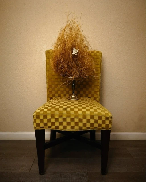 Still life photo of a yellow checkerboard chair with dried floral arrangement in a vintage metal candle holder against a cream wall.