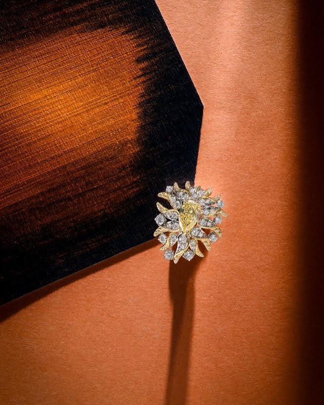 Close-up of an ornate gold ring with diamonds and a yellow gemstone set against a copper-colored surface with a dark painted corner accent.