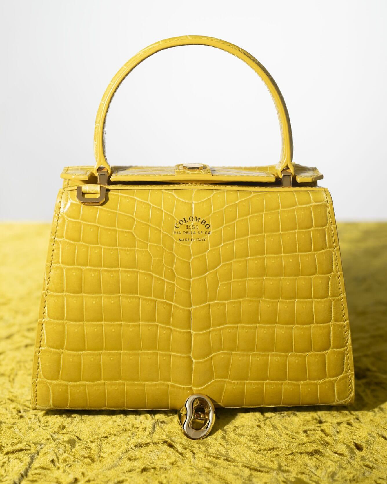Eye-level studio shot of a yellow alligator-texture handbag with a top handle, paired with a silver ring, set against a textured yellow background.