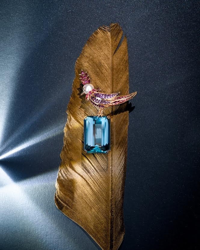 Close-up of a bird-shaped brooch with an aquamarine gemstone on a golden feather against a dark backdrop with light reflections.