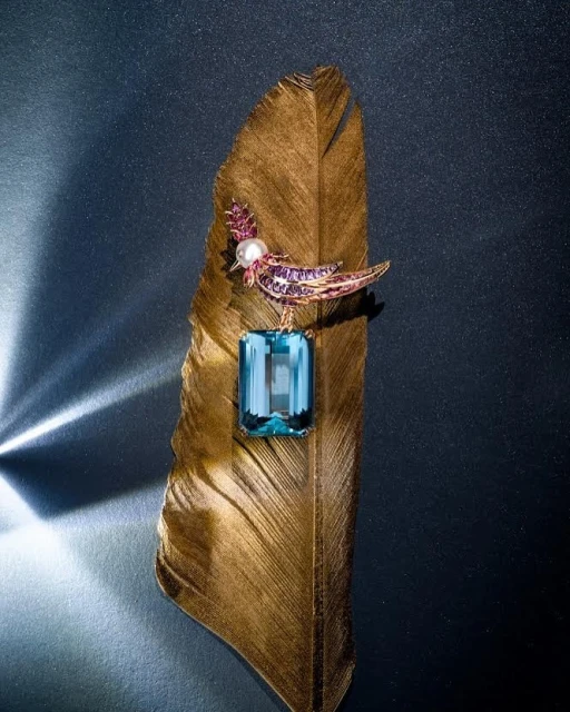 Close-up of a bird-shaped brooch with an aquamarine gemstone on a golden feather against a dark backdrop with light reflections.
