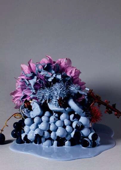 Painterly still life arrangement featuring grapes and pink-purple hued flowers covered in blue paint against a grey background.