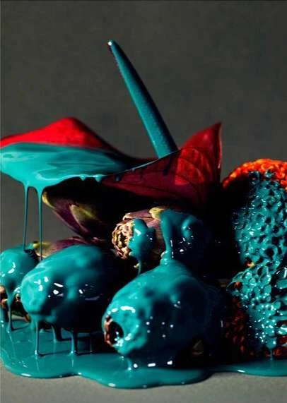 Still life of fruit and anthurium flowers covered in teal paint creating a vibrant and surreal visual, set against a grey backdrop.