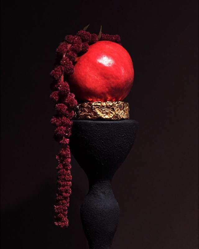 Still life photography of a red pomegranate fruit resting on a golden decorative pedestal, displayed on a black sculptural stand against a dark background.