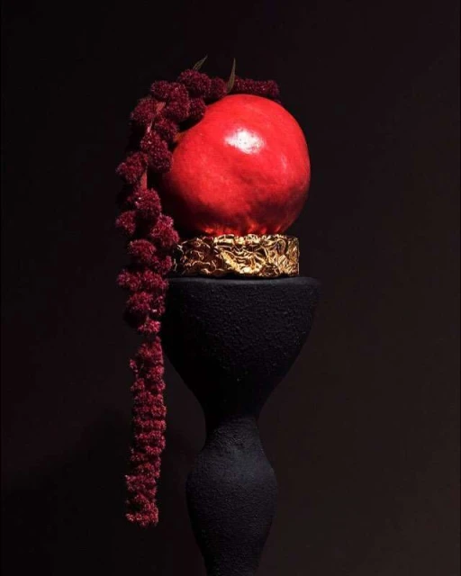 Still life photography of a red pomegranate fruit resting on a golden decorative pedestal, displayed on a black sculptural stand against a dark background.