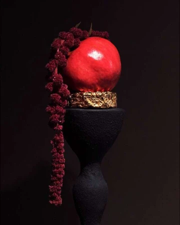 Still life photography of a red pomegranate fruit resting on a golden decorative pedestal, displayed on a black sculptural stand against a dark background.
