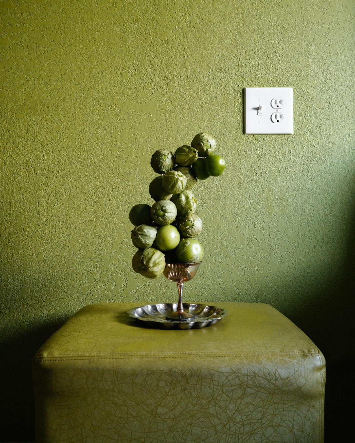 Still life of tomatillos in a silver cup on an ottoman against a green wall with an electrical outlet.