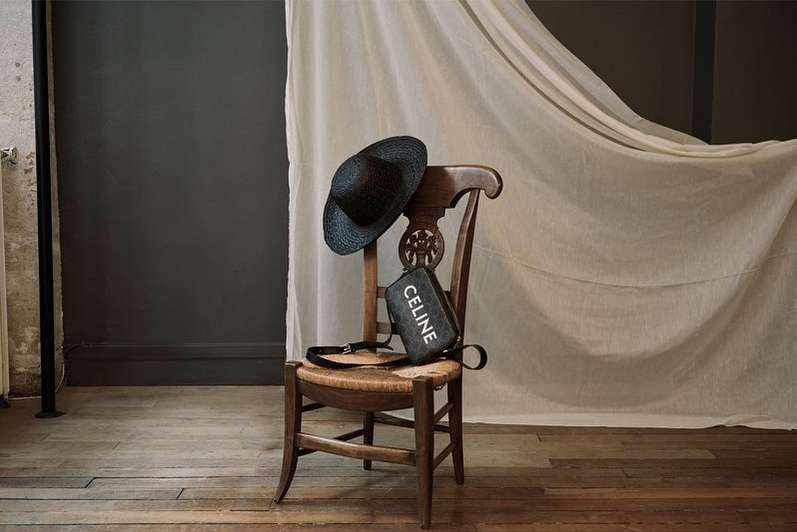 Still life of a Celine purse and black hat sitting on a rustic wooden chair against a neutral backdrop with dark gray wall.