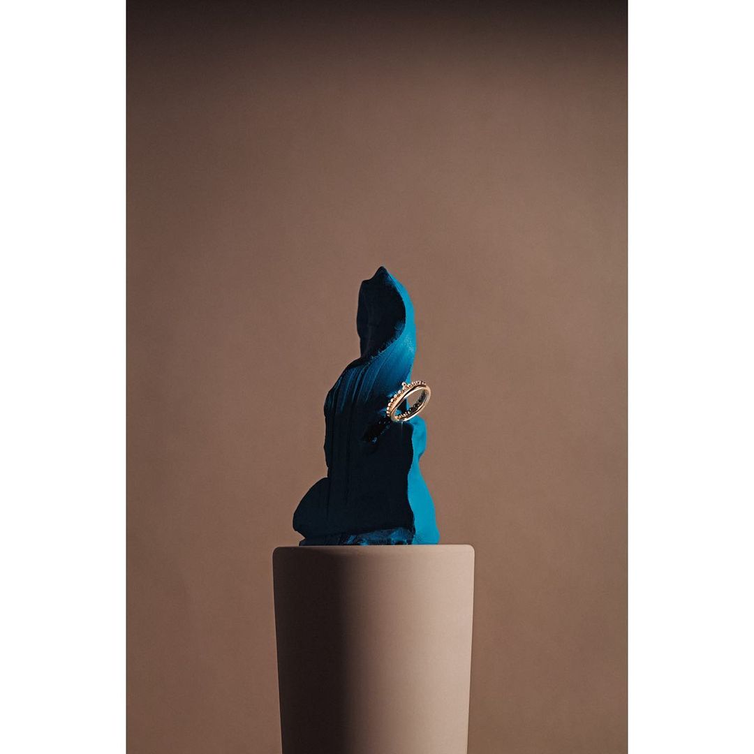 Close-up studio shot of ring on a teal sculpted form atop a cylindrical pedestal, neutral background, soft lighting.