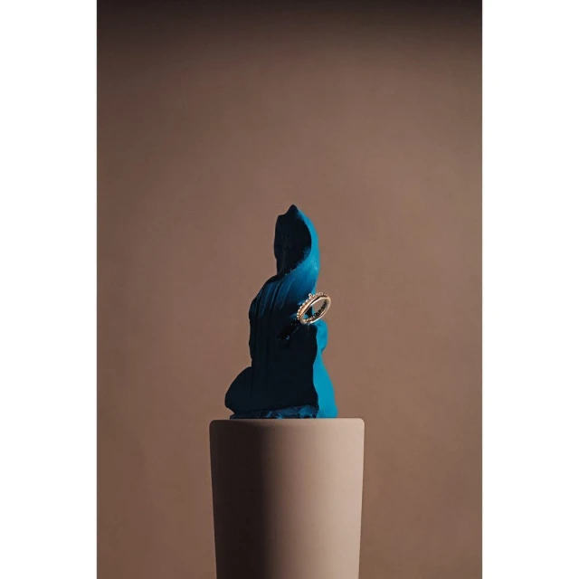 Close-up studio shot of ring on a teal sculpted form atop a cylindrical pedestal, neutral background, soft lighting.
