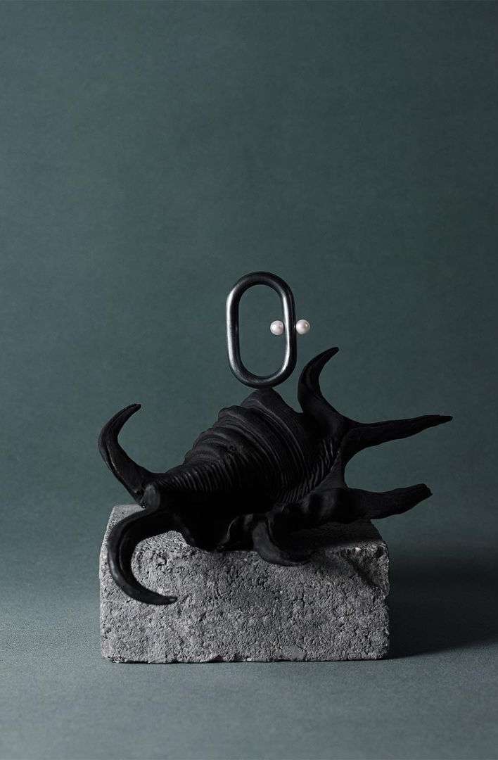 Abstract still life with a black seashell on a gray stone block, featuring a metal loop and two white pearls, against a teal background.