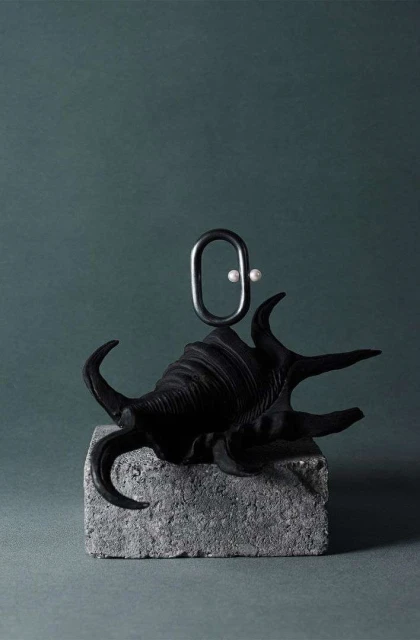Abstract still life with a black seashell on a gray stone block, featuring a metal loop and two white pearls, against a teal background.