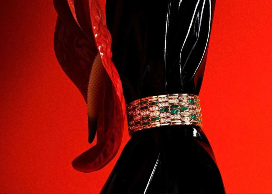 Elegant gold bracelet with green gemstones and diamonds on black glossy surface next to a red calla lily against a solid red backdrop.