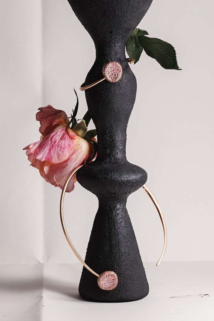 Black textured vase with gold jewelry and pink rose. Modern studio shot, emphasizing texture and color contrast. Minimalist composition.