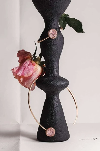 Black textured vase with gold jewelry and pink rose. Modern studio shot, emphasizing texture and color contrast. Minimalist composition.