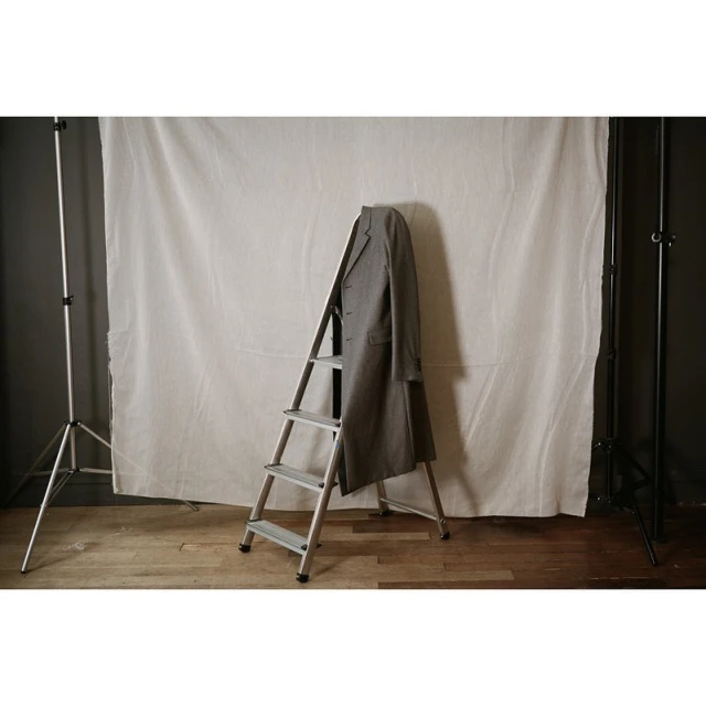 Grey wool coat draped on a silver step ladder in a photo studio with white backdrop and wooden floor. Fashion still life.