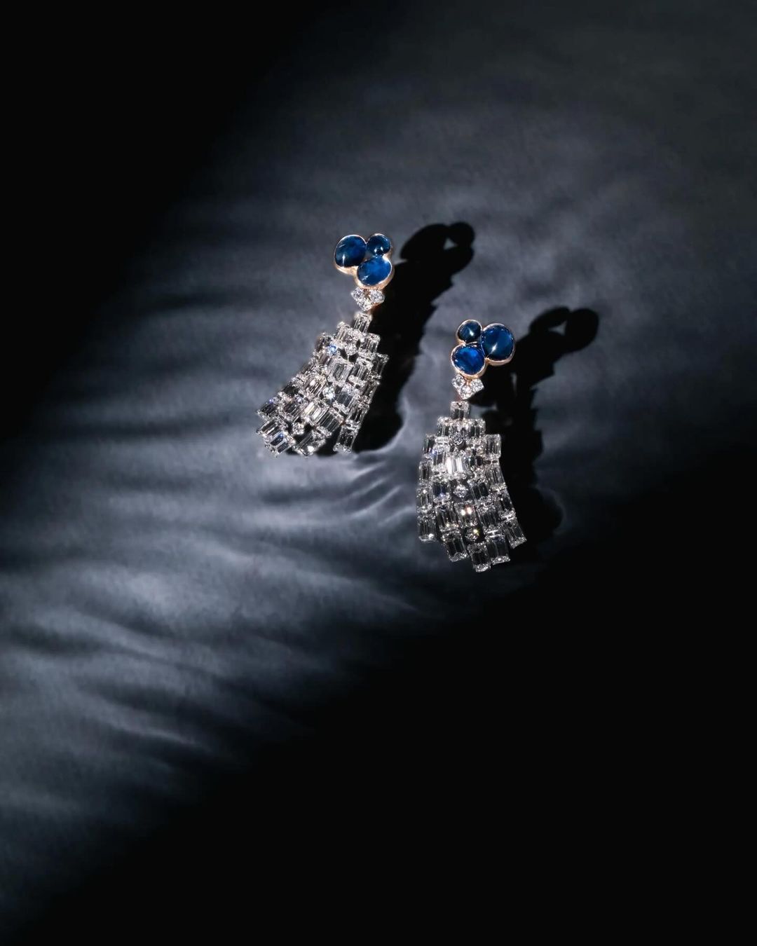 Elegant chandelier earrings with blue stones and geometric silver details on a dark reflective surface in a studio lighting setup.