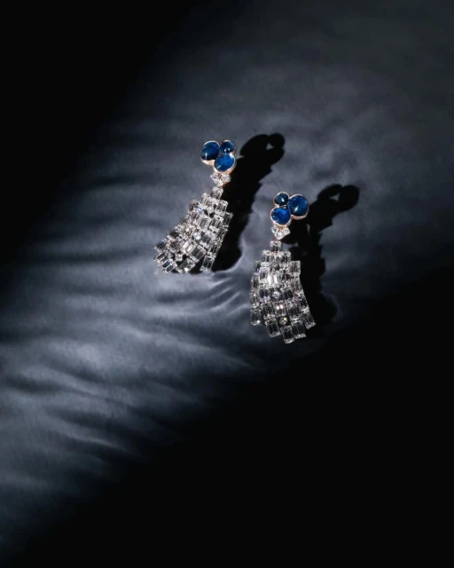 Elegant chandelier earrings with blue stones and geometric silver details on a dark reflective surface in a studio lighting setup.