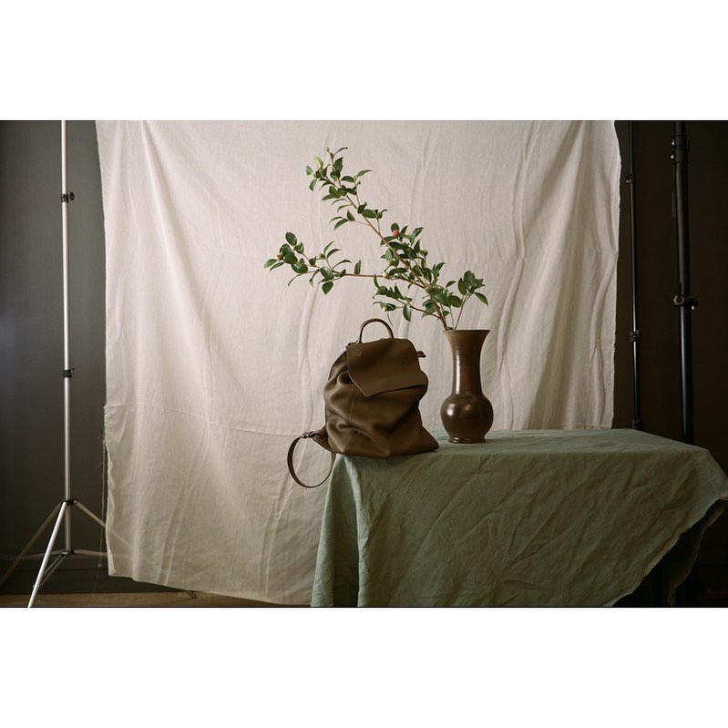 Still life of a brown leather backpack on a table with a vase and green foliage against a white backdrop in a photo studio.