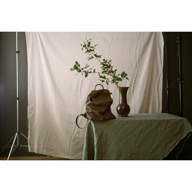 Still life of a brown leather backpack on a table with a vase and green foliage against a white backdrop in a photo studio.