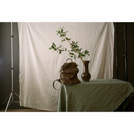 Still life of a brown leather backpack on a table with a vase and green foliage against a white backdrop in a photo studio.