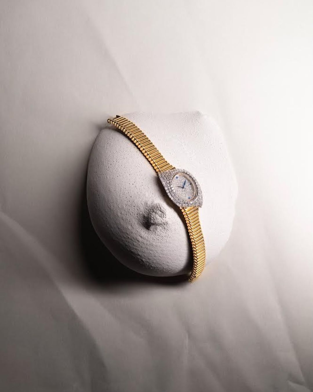 Elegant gold and diamond watch resting on a textured, abstract white bust against a soft, neutral-toned fabric backdrop.