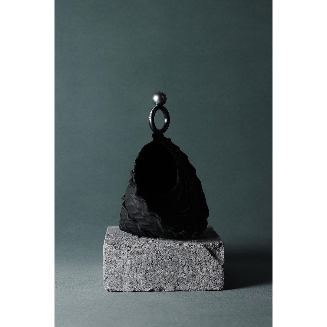Still life studio photograph features a dark ring with a pearl on top of a textured black stone, sitting on a gray concrete block.