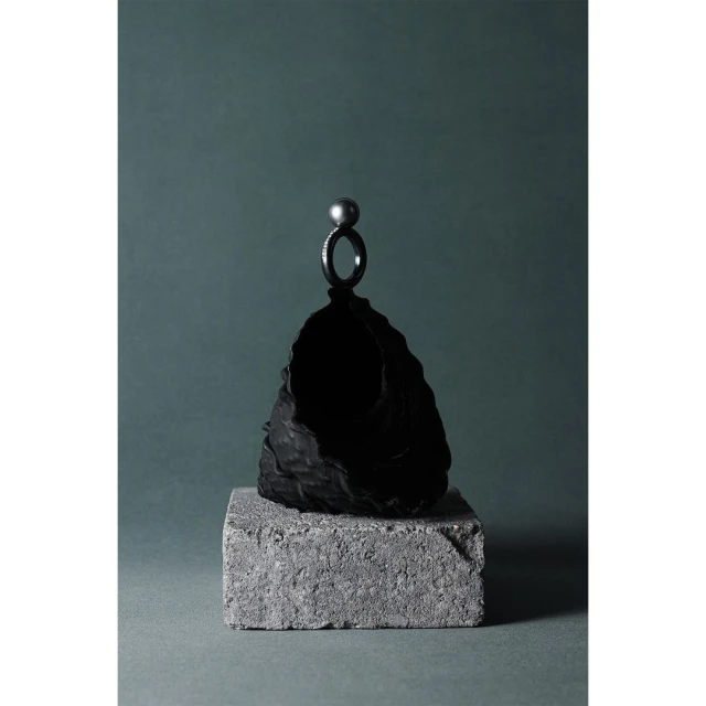Still life studio photograph features a dark ring with a pearl on top of a textured black stone, sitting on a gray concrete block.