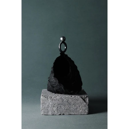 Still life studio photograph features a dark ring with a pearl on top of a textured black stone, sitting on a gray concrete block.