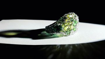 Close-up studio shot of a gem-studded ring featuring green and yellow gems set on a white surface against a dark background.