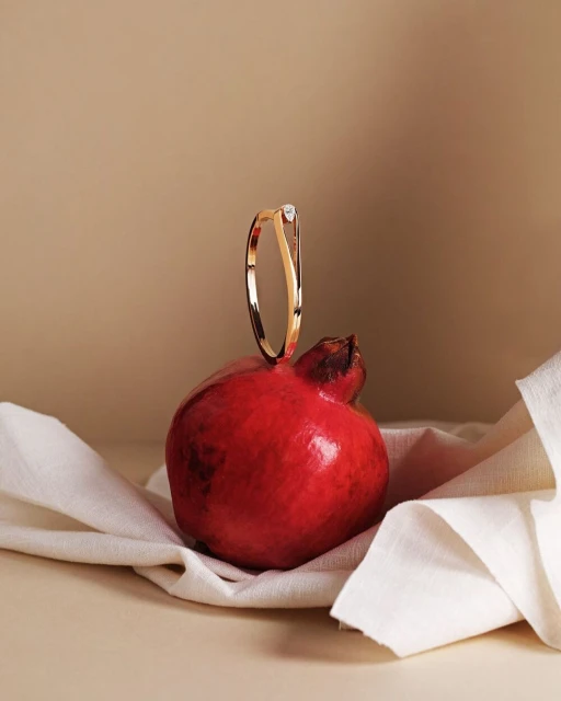 Close-up shot of a gold ring with a clear gemstone, balanced on a ripe pomegranate, against a gradient tan background. Jewelry product shot.