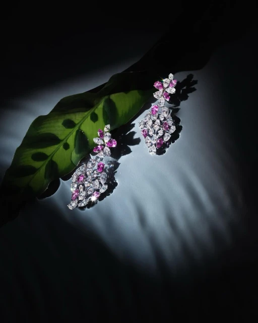Elegant floral drop earrings featuring pink and white gemstones displayed on a vibrant green leaf, spotlighted against a dark background.