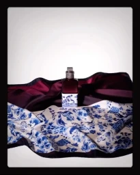 A perfume bottle standing on a patterned blue and white silk scarf with a dark burgundy draped fabric background.