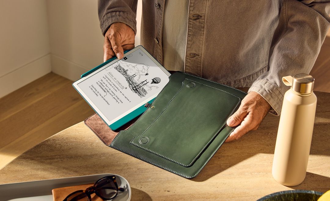 Person holding an open tablet with a drawing on screen and a green folio case on a wood table with a water bottle nearby.