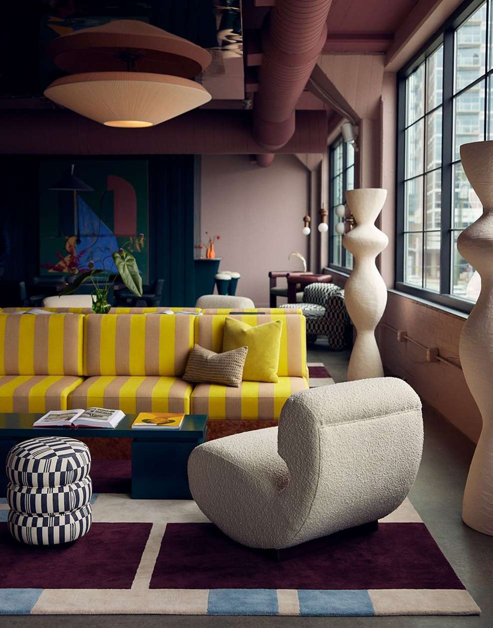 Living room interior with striped yellow sofa, beige armchair, patterned ottoman, dark blue table, sculpture, and large windows.