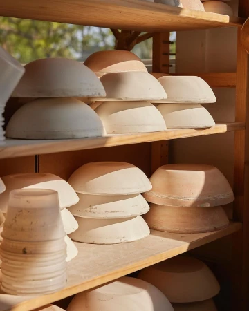 Stacks of handmade ceramic bowls on wooden shelves in a brightly lit pottery workshop. Bowls are beige and arranged neatly. 