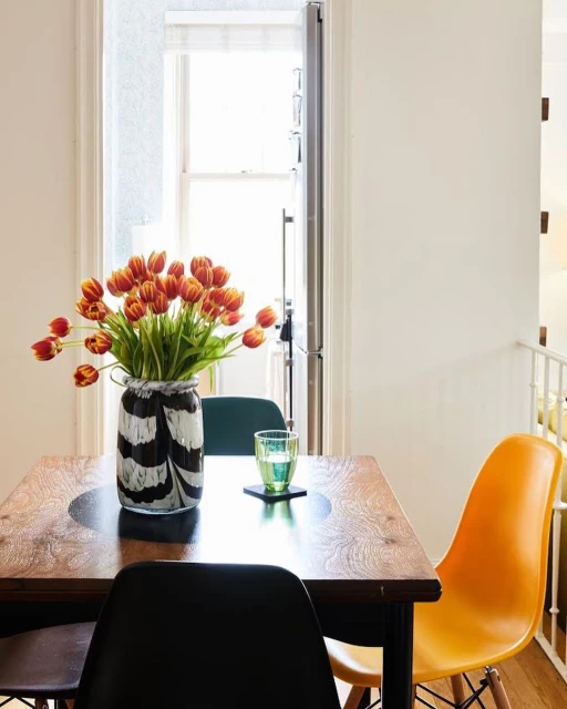 A bouquet of red and yellow tulips in a black and white vase sits on a wooden table with colorful chairs in a bright indoor setting.