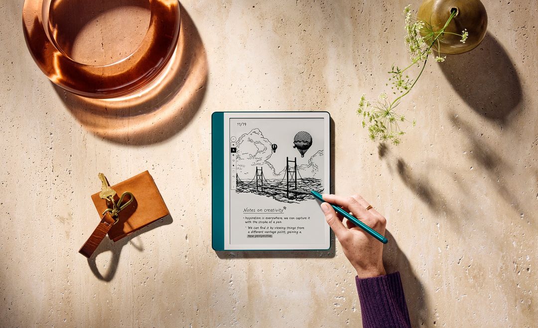 E-reader on a travertine surface with a sketch of a bridge and hot air balloon, surrounded by accessories, stylus and flowers.