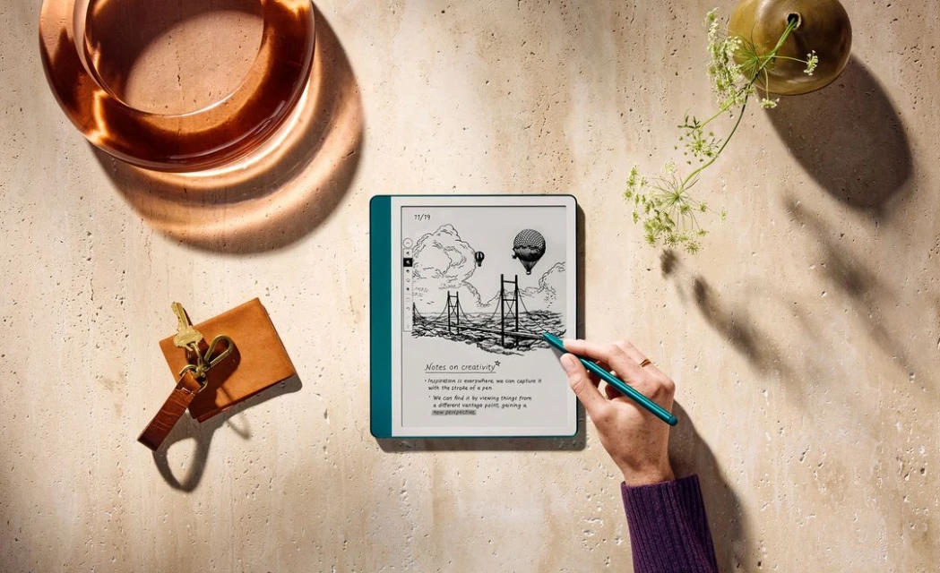 E-reader on a travertine surface with a sketch of a bridge and hot air balloon, surrounded by accessories, stylus and flowers.