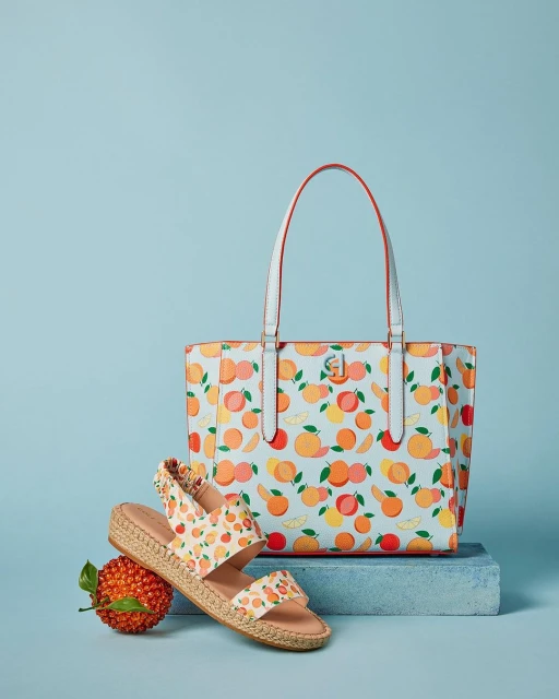 Still life of a tote bag with white handles and a fruit pattern, plus sandals with a similar pattern, on a light blue background.