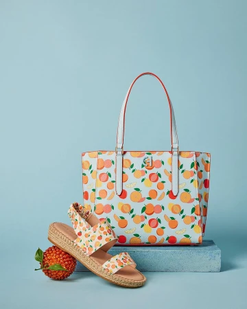 Still life of a tote bag with white handles and a fruit pattern, plus sandals with a similar pattern, on a light blue background.