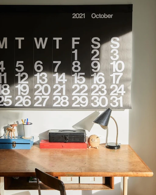 A warm-toned interior scene features a wooden desk, a black desk lamp, and a 2021 October calendar hanging above it with natural light.