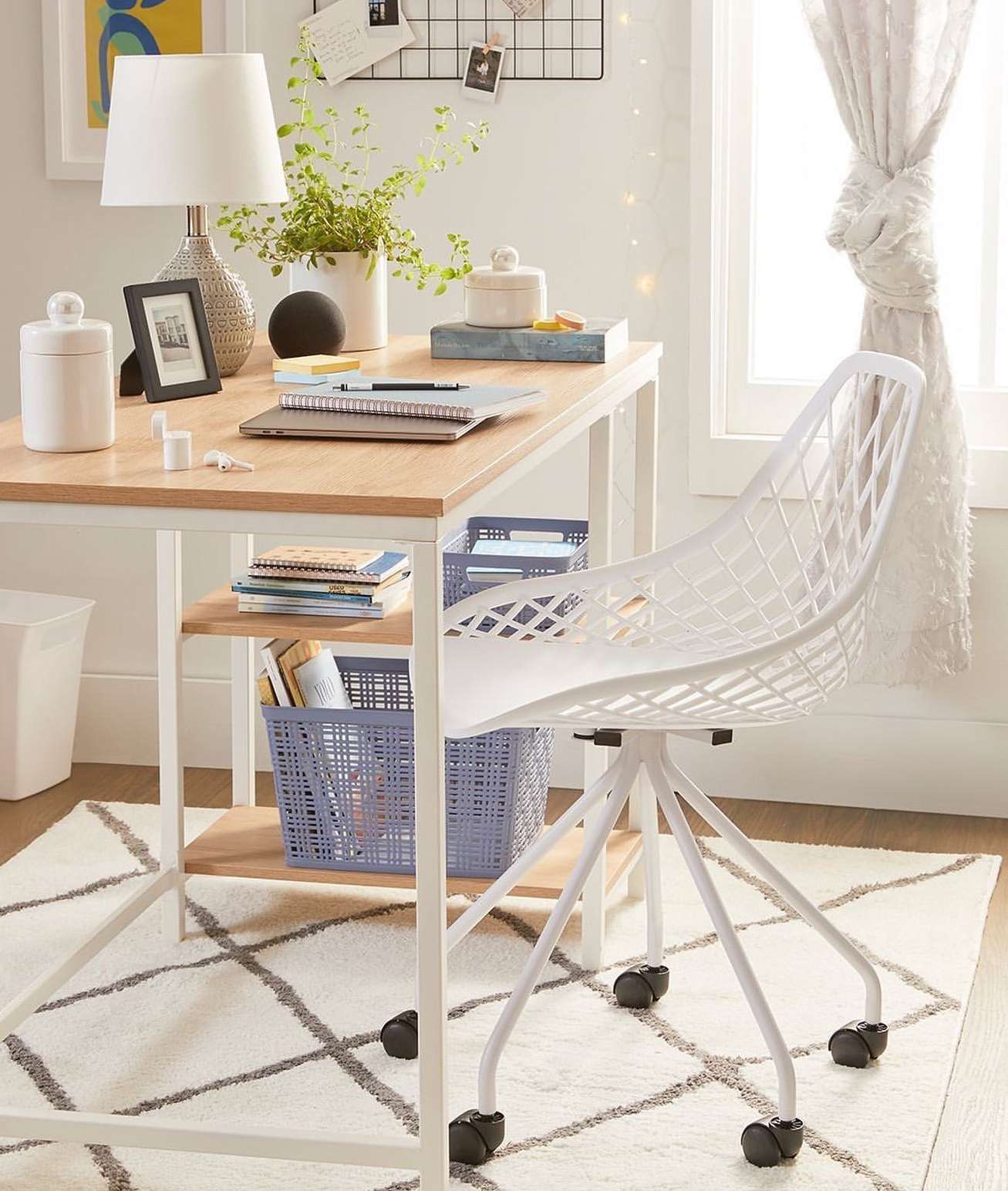 Home office setup with a light wood desk, white chair, lamp, and decor, set against a white and gray patterned rug and natural light.